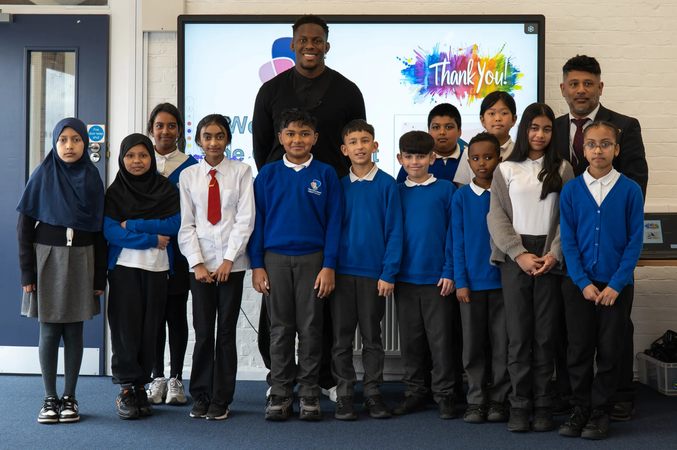 Maro Itoje and a school worker stand with a group of students in a classroom.