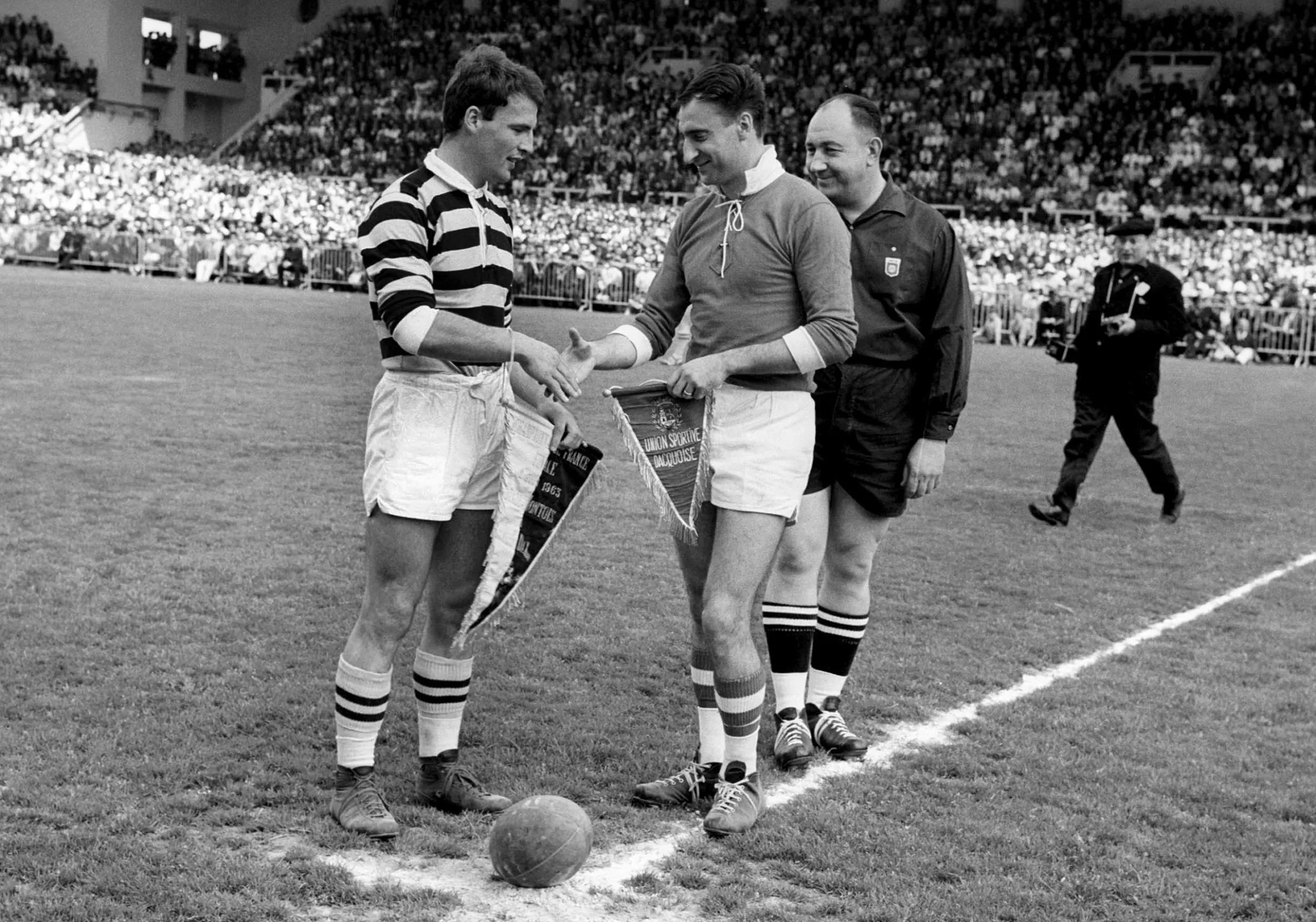 A black and white photograph of two rugby players holding hands on the pitch with an official standing beside them and the crowd behind.