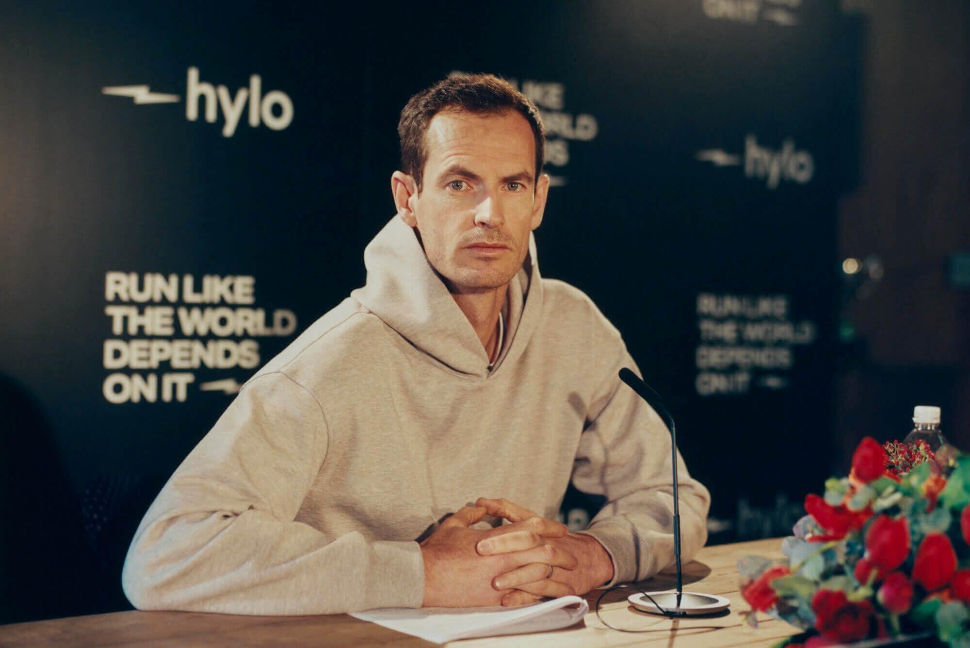 Andy Murray sits wearing a beige hoodie in front of a backdrop with the Hylo logo and slogan on it.
